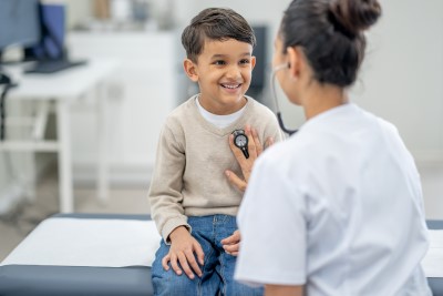 A young boy sits up on an exam table in a doctors office during a routine check-up. His doctor is seated in front of him and listening to his heart with her stethoscope.